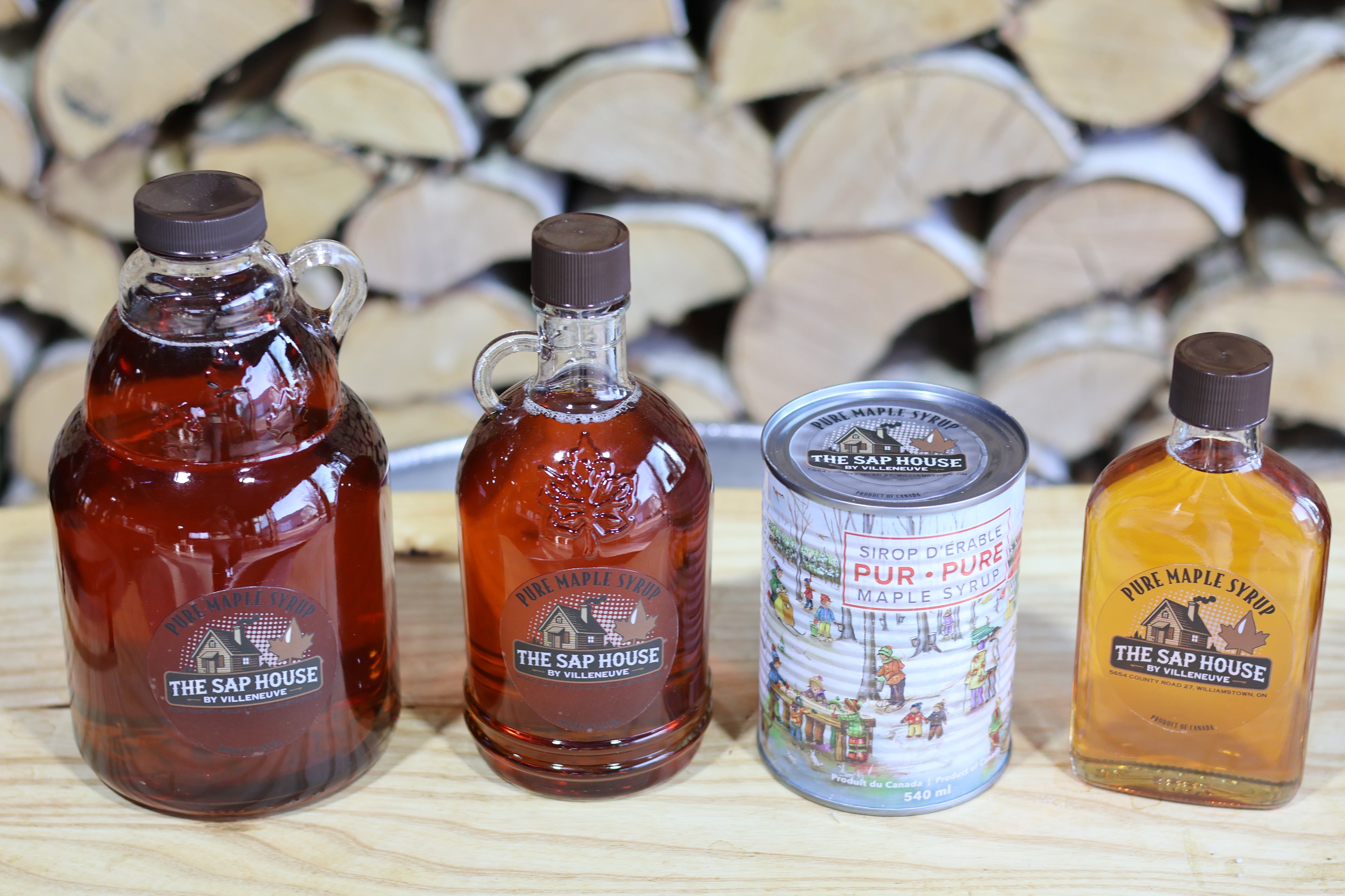 Three bottles and a can of The Sap House maple syrup on a wooden surface with stacked firewood in the background.