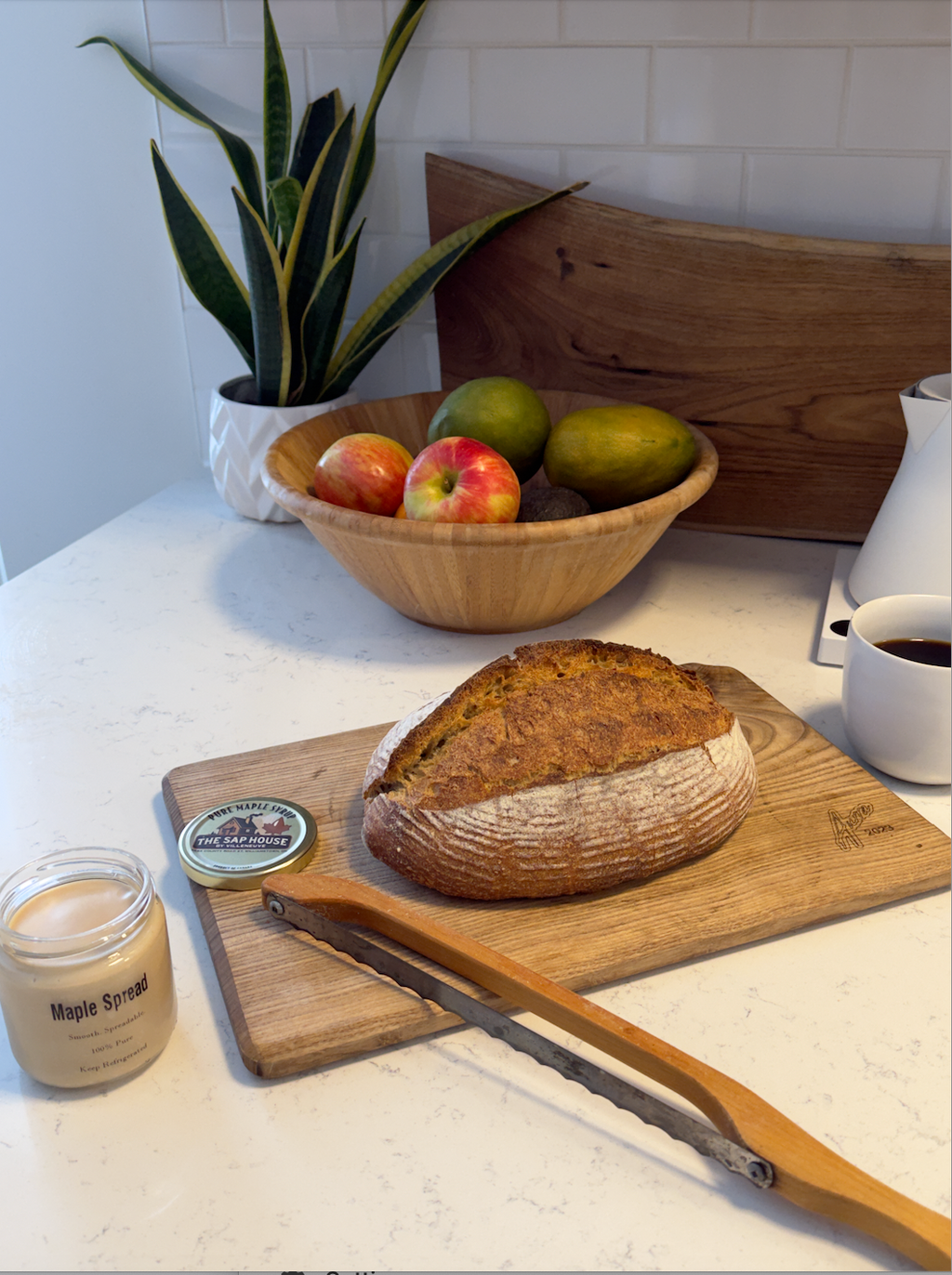 maple spread, a loaf of bread and a bread knife on a cutting board with a fruit basket in the back.