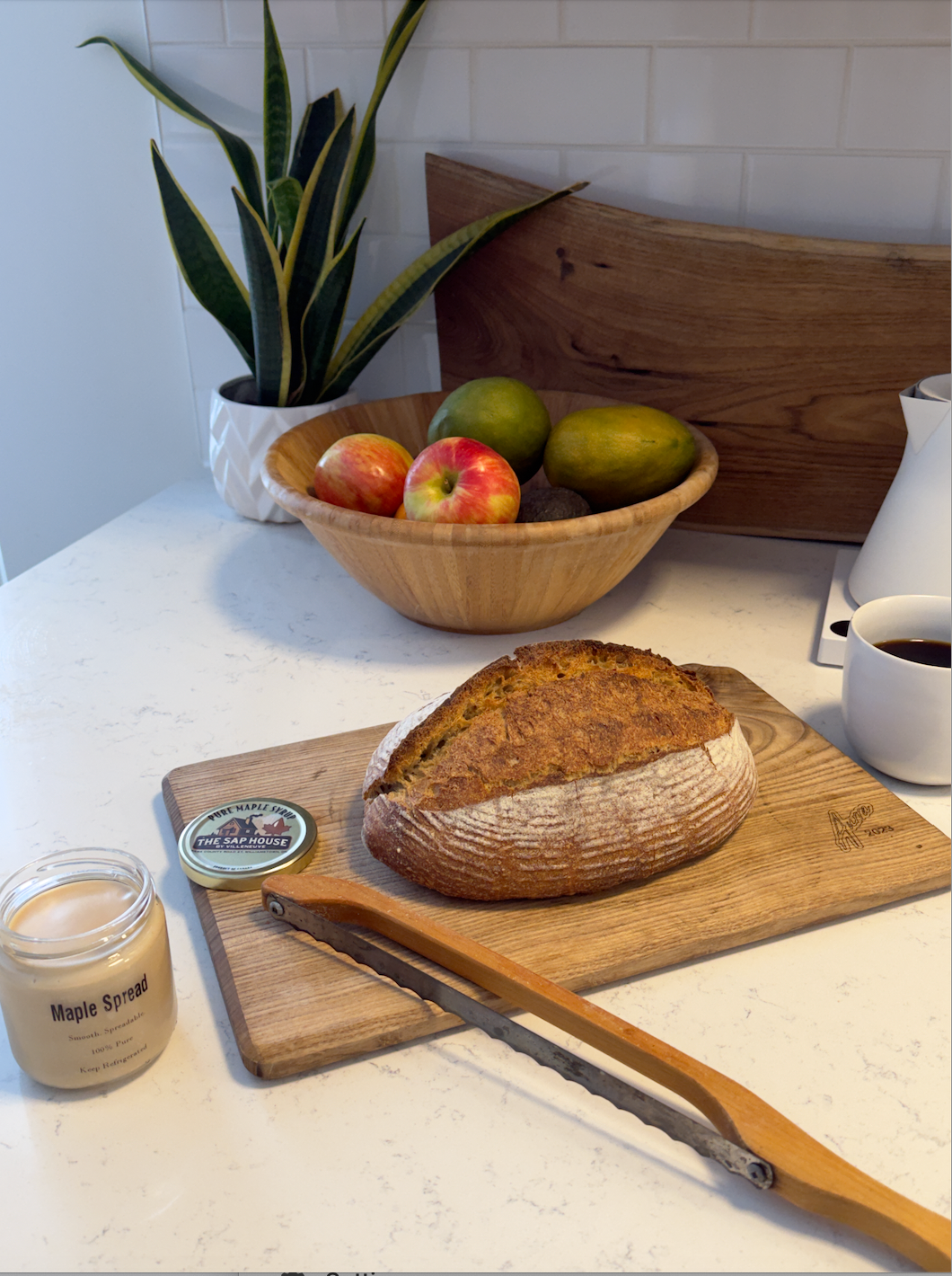 maple spread, a loaf of bread and a bread knife on a cutting board with a fruit basket in the back.