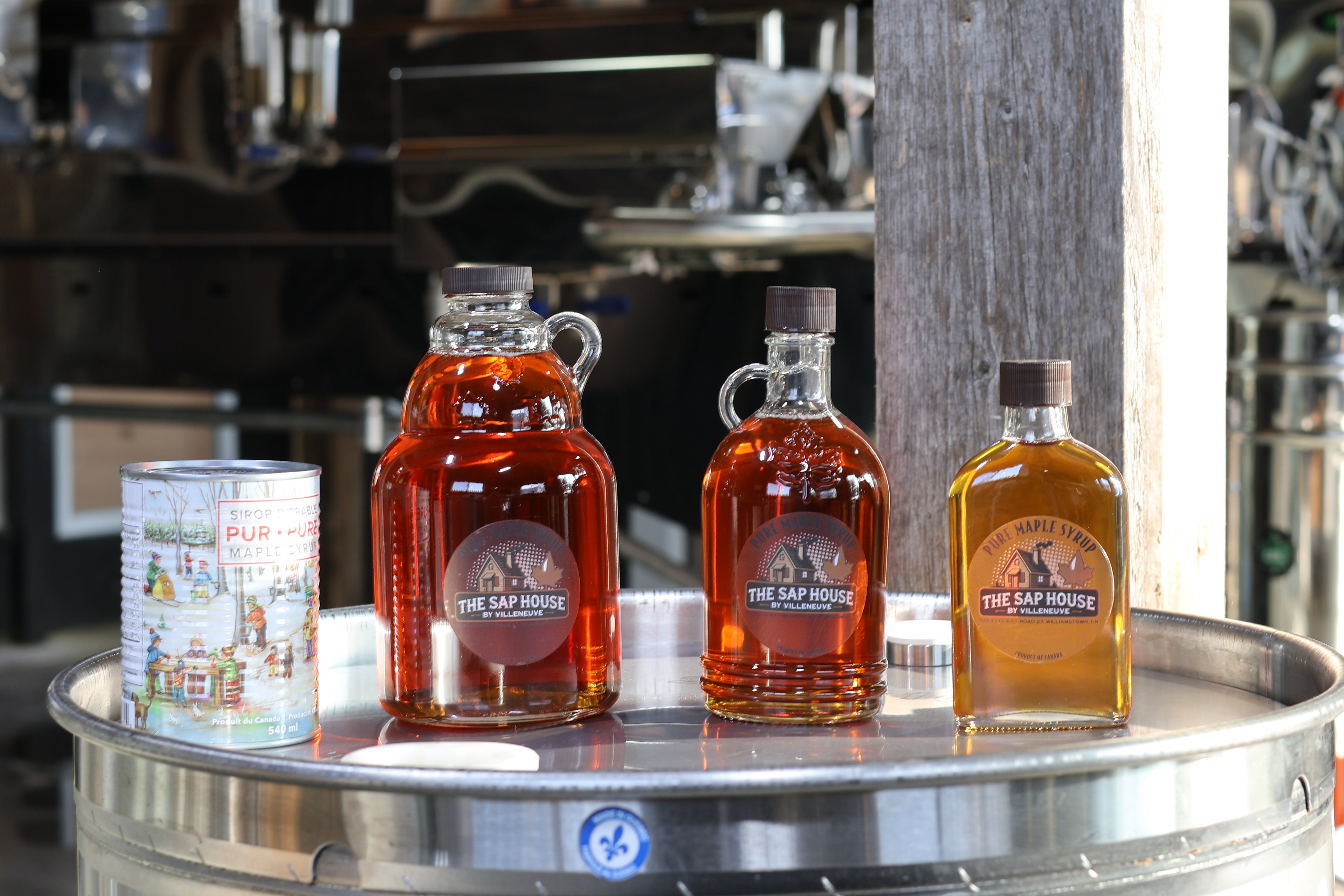 Three bottles and a can of The Sap House maple syrup on a metal surface with a blurred background.